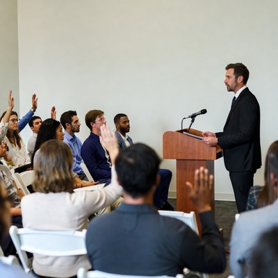 Man speaking at podium with audience raising hands