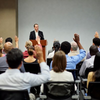 Man speaking at podium with audience raising hands